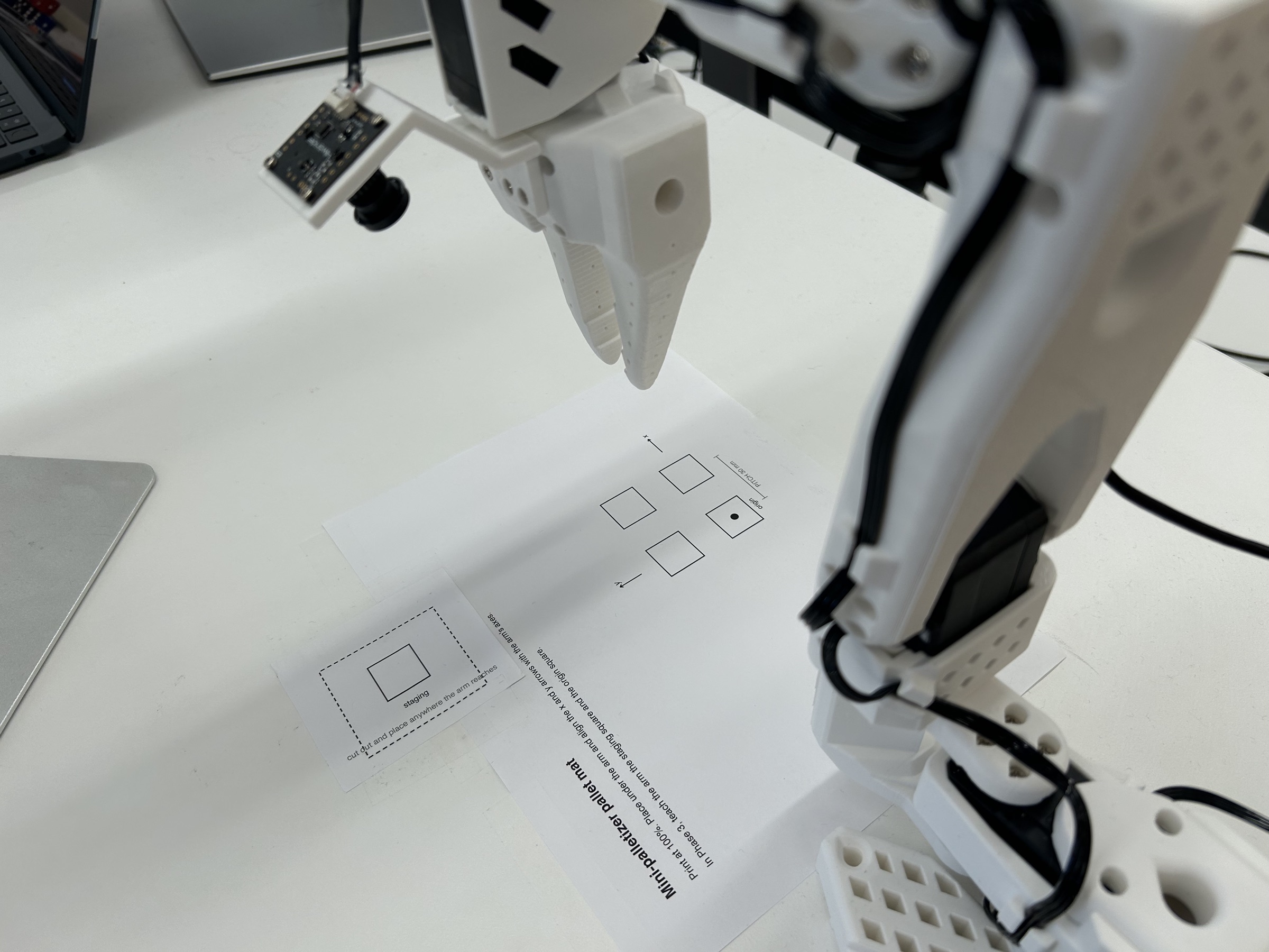 Looking out from behind the SO-ARM101's base at the printed pallet mat and staging square taped to the desk. The mat's x arrow points straight out, away from the base, and its y arrow points to the arm's left; the origin square, marked with a dot, is the cell nearest the base.